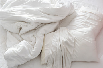 Top view of an unmade bed in a bedroom with crumpled bed sheet, a blanket, a white shower towel and two pillows after waking up in the morning. Not having bedclothes neatly arranged for sleep in.