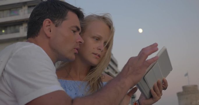 Steadicam shot of young man and woman using tablet computer on waterfront with hotels and walking people