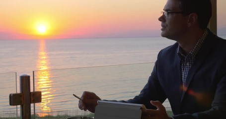 Man using tablet computer to work on business. He sitting in cafe with view to the oversea sunset and smoking - Powered by Adobe