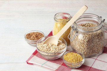 various cereals in glass jars and wooden spoon with glass bowls on a napkin on a white wooden table