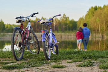 Fototapeta premium Two bicycles in the foreground and mother with daughter standing at the lake shore in the blurred background 