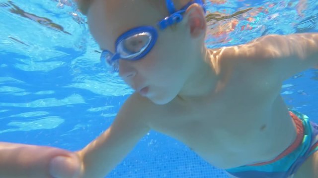 Slow motion clip of a little boy swimming in goggles under clean blue water  in the pool. Someone helping him swim with the hands