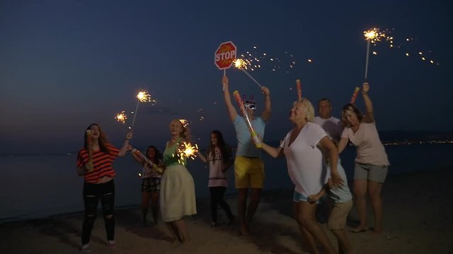 Slow motion clip of funny people dancing and having fun on the beach at night. They have sparklers and firecrackers in arms