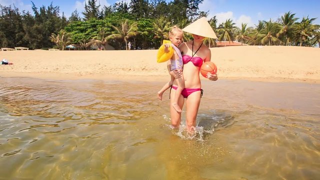Mother in Hat Carries in Arms Daughter into Sea against Palms