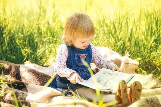 Child reading book in park. Portrait of cute little girl sitting on picnic blanket holding big book in hands. Little girl looking at pictures and learning to read.