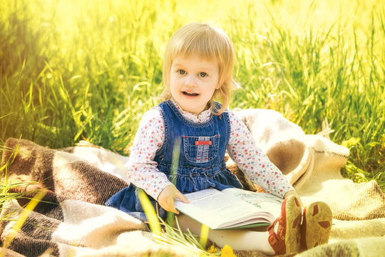 Child reading book in park. Portrait of cute little girl sitting on picnic blanket holding big book in hands. Little girl looking at pictures and learning to read.
