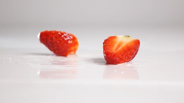 Fresh tasty red sliced strawberry fly and breaking on two halfs on white wet surface with explosive splash and spray and bouncing. Shot with high speed camera in slow motion mode. Gray background