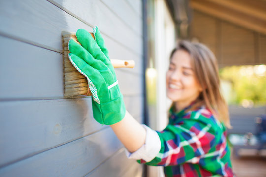 Cheerful woman applying protective varnish or paint on wooden house tongue and groove cladding elevation wall. Focus on hand with brush. House improvement diy concept.