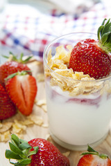 Homemade healthy dessert in a glass with yogurt, fresh strawberry and corn flakes for breakfast with fresh white flowers and checkered napkin on wooden table, closeup