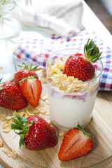 Homemade healthy dessert in a glass with yogurt, fresh strawberry and corn flakes for breakfast with fresh white flowers and checkered napkin on wooden table, closeup