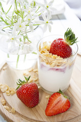 Homemade healthy dessert in a glass with yogurt, fresh strawberry and corn flakes for breakfast with fresh white flowers on wooden table, closeup