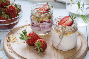 Homemade healthy dessert in a glass with yogurt, fresh strawberry and corn flakes for breakfast with fresh white flowers on wooden table, closeup