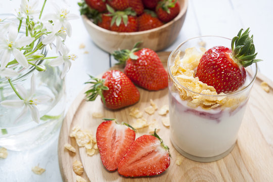 Homemade healthy dessert in a glass with yogurt, fresh strawberry and corn flakes for breakfast with fresh white flowers on wooden table, closeup