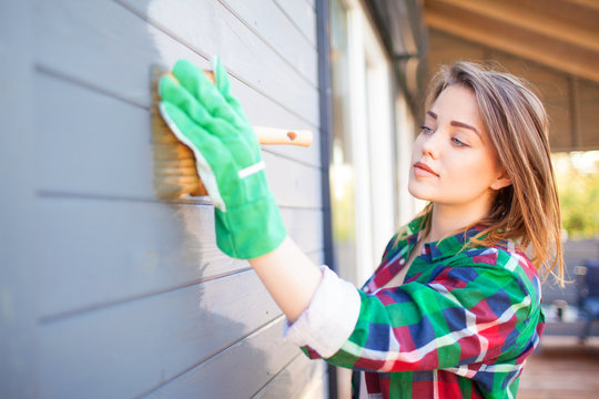 Young woman applying protective varnish or paint on wooden house tongue and groove cladding elevation wall. House improvement diy concept.