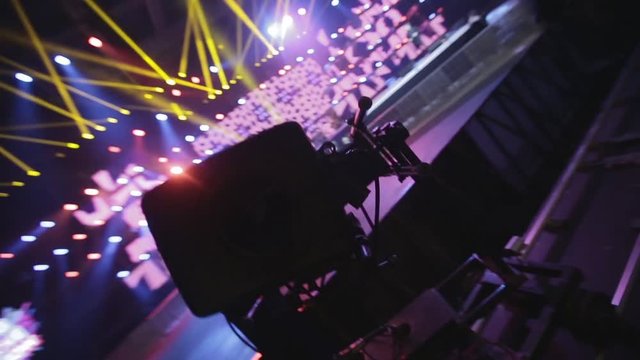 Concert backstage. Close up of video camera in action with pink, yellow and purple spotlights and the stage in the background.