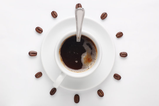Coffee cup with spoon on saucer and coffee beans against white background forming clock dial viewed from top