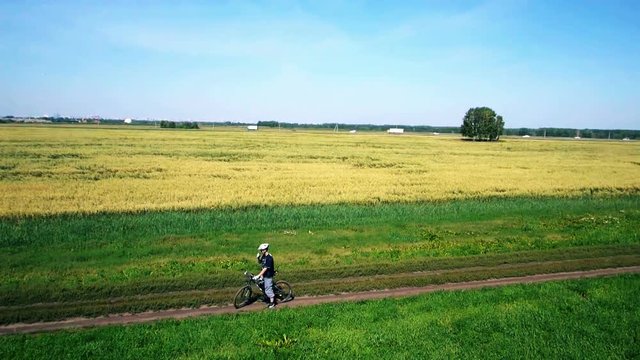 AERIAL: Young man cycling on bicycle at rural road through green and yellow summer meadow field. During sunny day.
