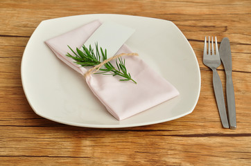 A simple place setting consisting of a plate and a knife and fork with a napkin that is tied together an empty name card and a piece of a plant
