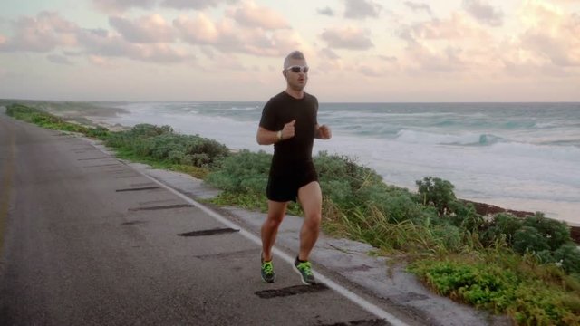 man athlete runner in sport dress and sunglasses working out running at the sea slow motion steadicam