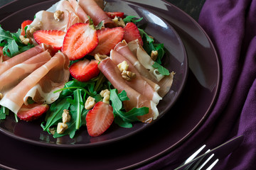 Fresh salad with strawberry, arugula and prosciutto on purple plate. Low key
