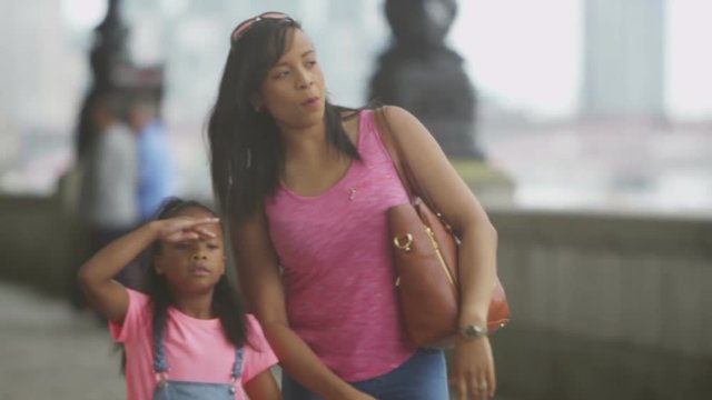 Happy African American mother & daughter sightseeing in London, walking along path beside the river Thames. 