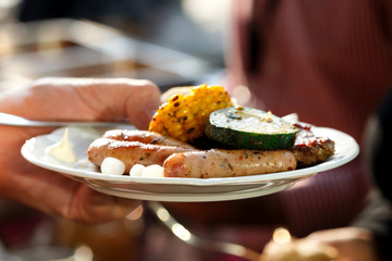 Hand holding a plate of barbecued meat, sausages and vegetables.