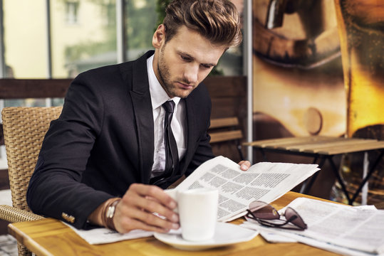 A man in a suit reading a newspaper and drinking coffee.