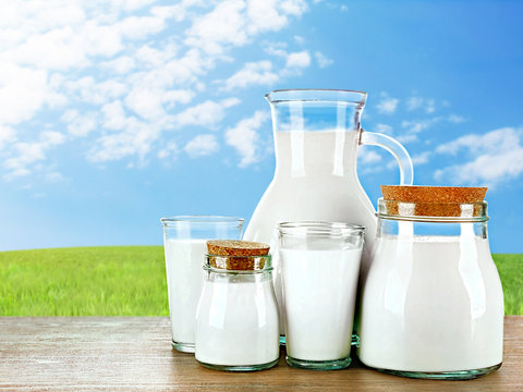 Pitcher, jars and glasses of milk on wooden table against green field and blue sky background