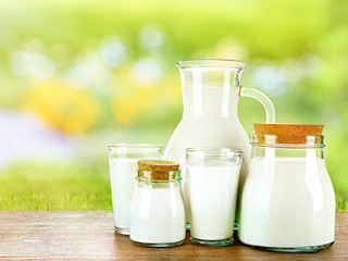 Pitcher, jars and glasses of milk on wooden table against blurred green background