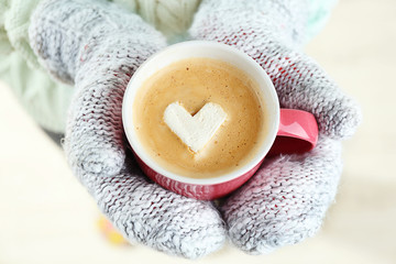 Female hands in warm mittens holding cup of hot cappuccino with heart marshmallow, close up