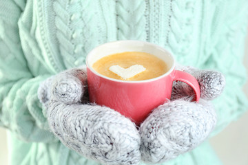 Female hands in warm mittens holding cup of hot cappuccino with heart marshmallow, close up