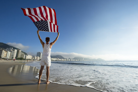Athlete in white outfit standing with American flag waving in the wind on the shore of Copacabana Beach, Rio de Janeiro, Brazil