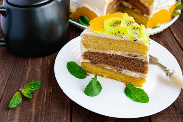 A piece of fruit cake (kiwi, orange, mint leaves) on a white plate on wooden background close-up and a cup of tea