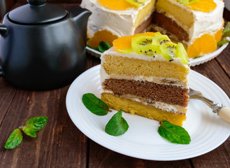 A piece of fruit cake (kiwi, orange, mint leaves) on a white plate on wooden background close-up and a cup of tea