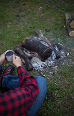 Man drinking coffee by the fire at a campsite on the river bank.