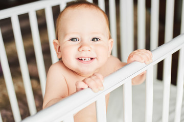 A year old baby is smiling in the crib and holds onto the side of the bed