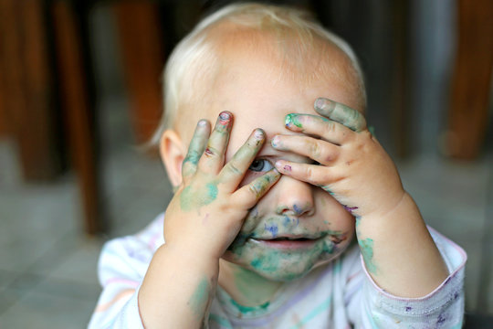 Funny one year old girl covering messy painted face with her dirty little hands