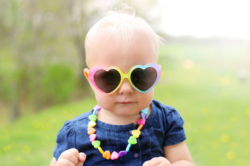 Cute one year old baby girl wearing heart shaped sunglasses and necklace outside on a sunny summer...