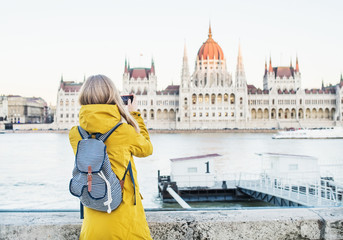 Fototapeta premium Young blondy woman tourist making photos of Parliament historic building with her phone in Budapest, Hungary