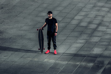 Stylish young hipster man standing with a longboard on a background of concrete tiles. Bearded man in bright sneakers, sunglasses and a stylish cap in the city urban landscape.