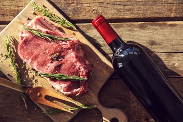 Crédence de cuisine en verre imprimé Viande Steak de porc cru avec bouteille de vin rouge, fourchette à viande et romarin sur fond de bois