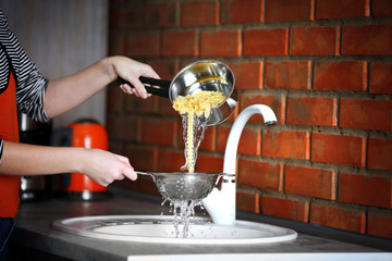 Female hands pouring water from boiled pasta over sink in the kitchen - obrazy, fototapety, plakaty