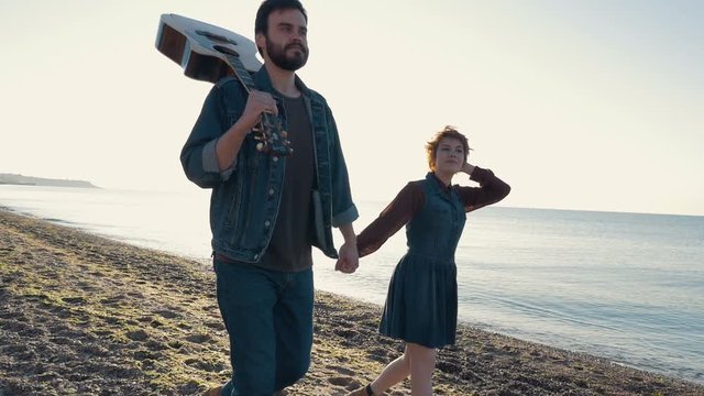 happy loving couple walking on beach during sunrise or sunset with guitar