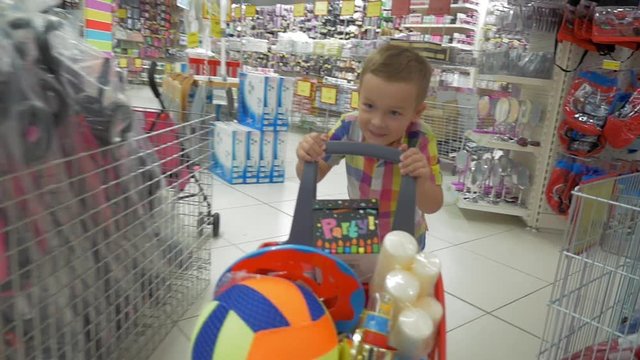 Slow motion steadicam clip of  a smiling boy in the mall going with a shopping cart full of party things. Scene in a big supermarket with a lot of different goods around