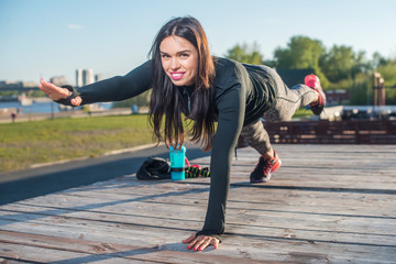 Fit woman doing raised leg plank yoga pilates exercises training her abs core muscles outdoors looking at camera