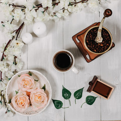 Morning coffee with coffee mill, beans, candles, green leaves sakura branch, vintage white tray, pink rose flowers on a white wooden table. Top view, flat lay