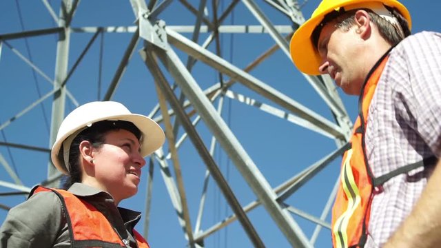 Close up low angle shot of a male and female electrical workers discussing the situation with a high tension power pole structure in the background.