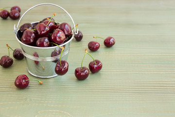 Bucket of cherries.In the frame on the left a small bucket with cherries, around scatterd cherries, on the right empty space on a light green background. Horizontal. Daylight.