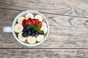 very delicious acai bowls with fresh fruit strawberry, blueberry, banana and peppermint leaves on top in cute white cup on the wooden table. this smoothie dessert is good for summer.