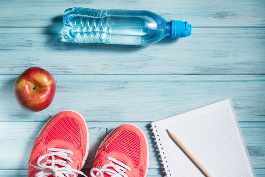 Fitness concept, pink sneakers, red apple, bottle of water and notebook with pencil on wooden background, top view